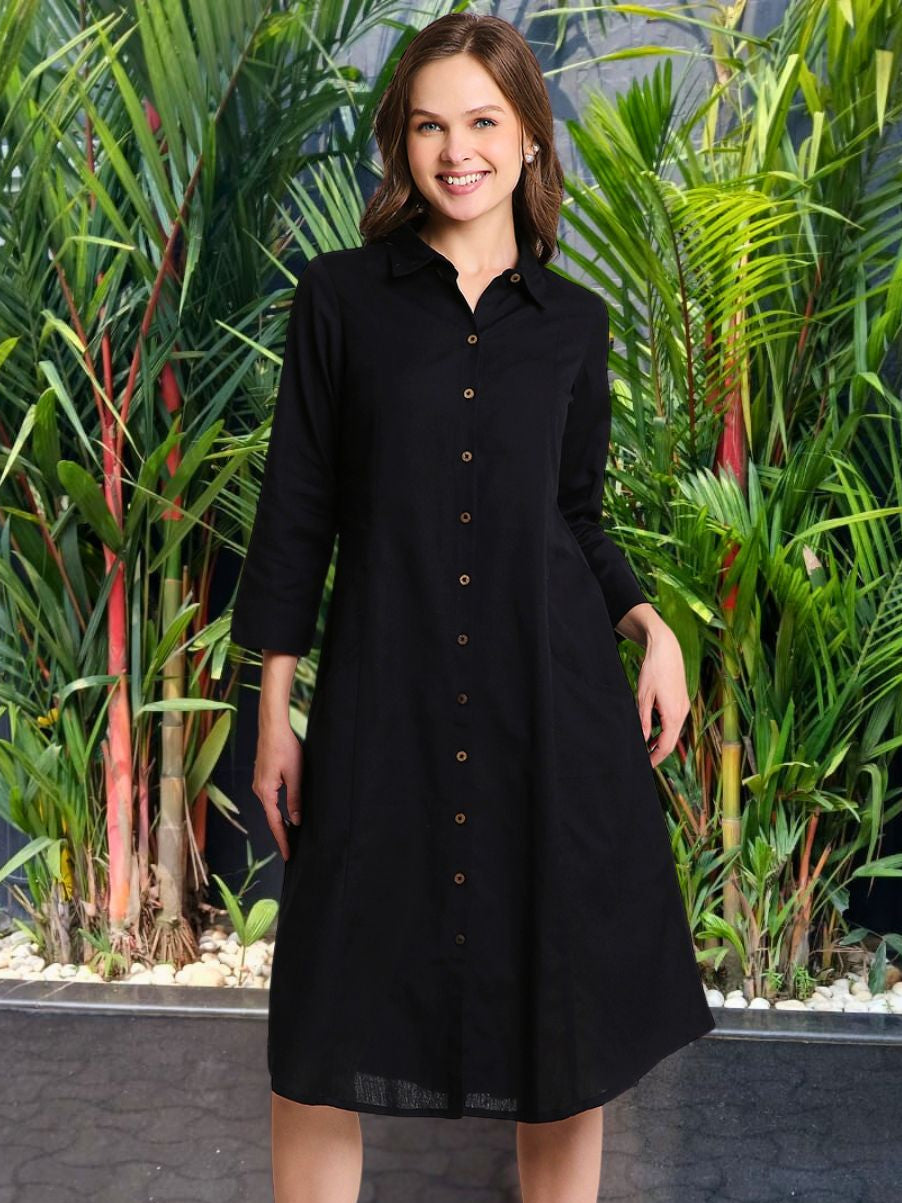 A woman standing in front of plants, wearing a black solid midi shirt dress with a classic shirt collar, 3/4th sleeves, and an A-line silhouette.